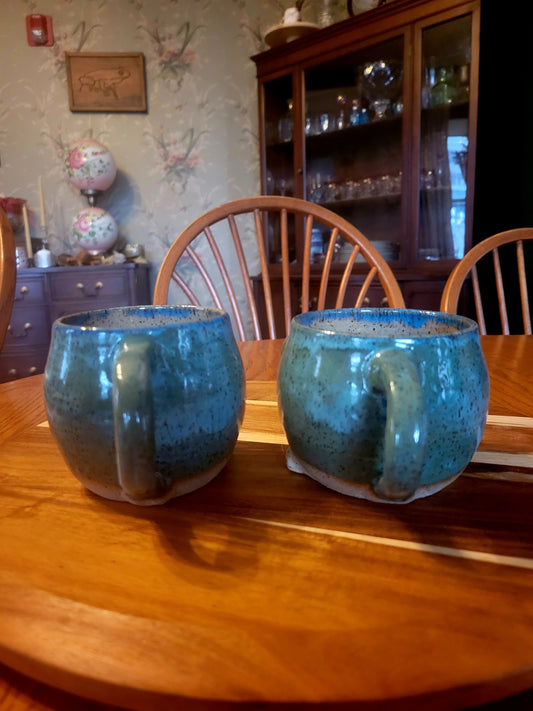 Two blue ceramic mugs on a wooden table with a background of a room with furniture and decor.