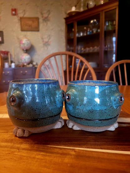 Two blue ceramic frog-shaped mugs on a wooden table with a dining room background.