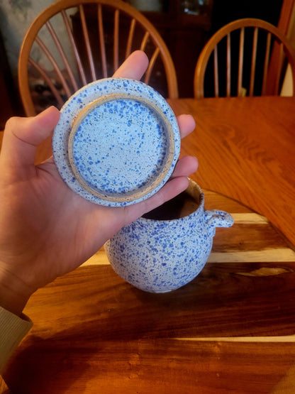Speckled ceramic pot and lid held by a hand on a wooden table.