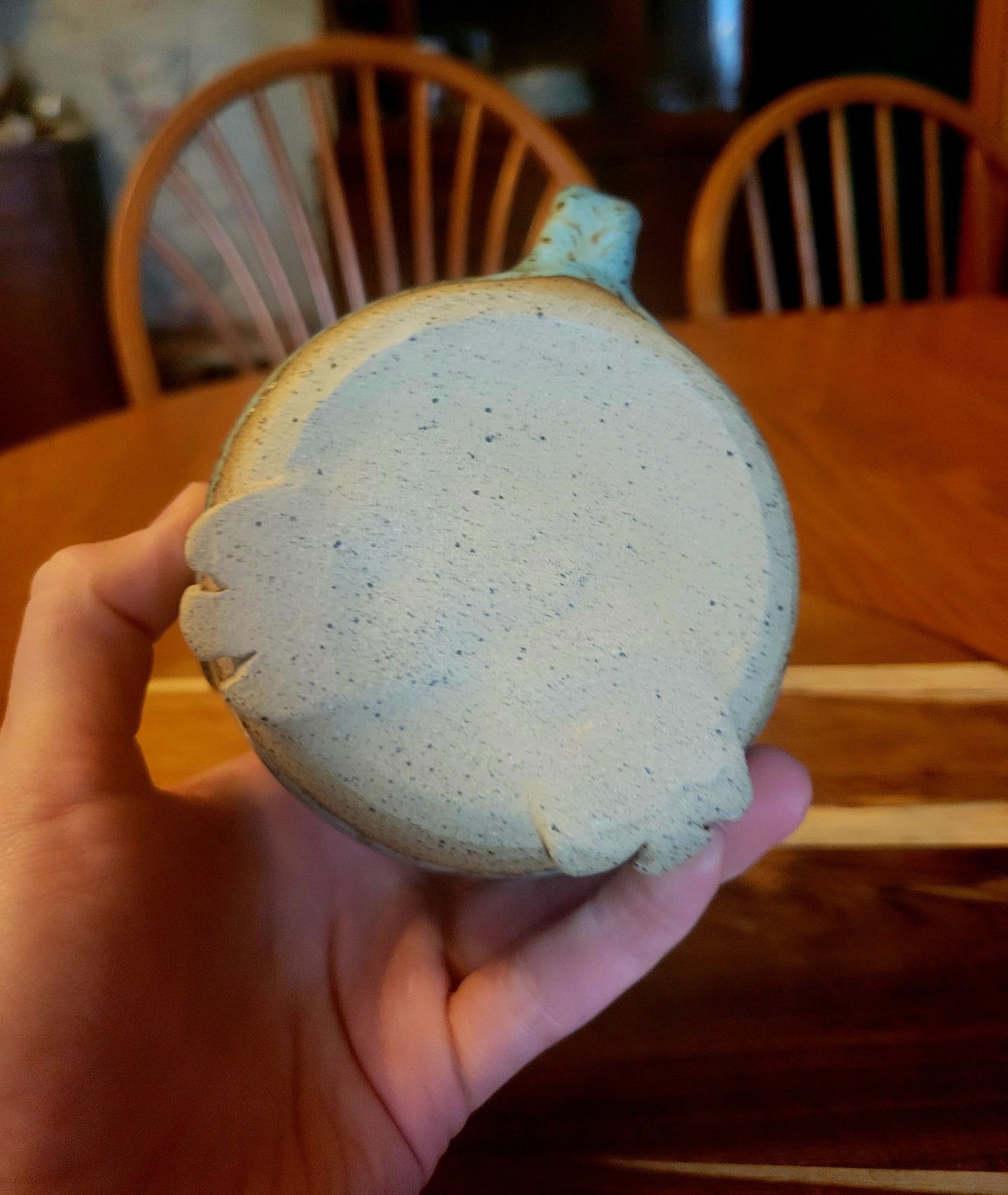 Hand holding a round, speckled ceramic mug with a wooden chair and table in the background.