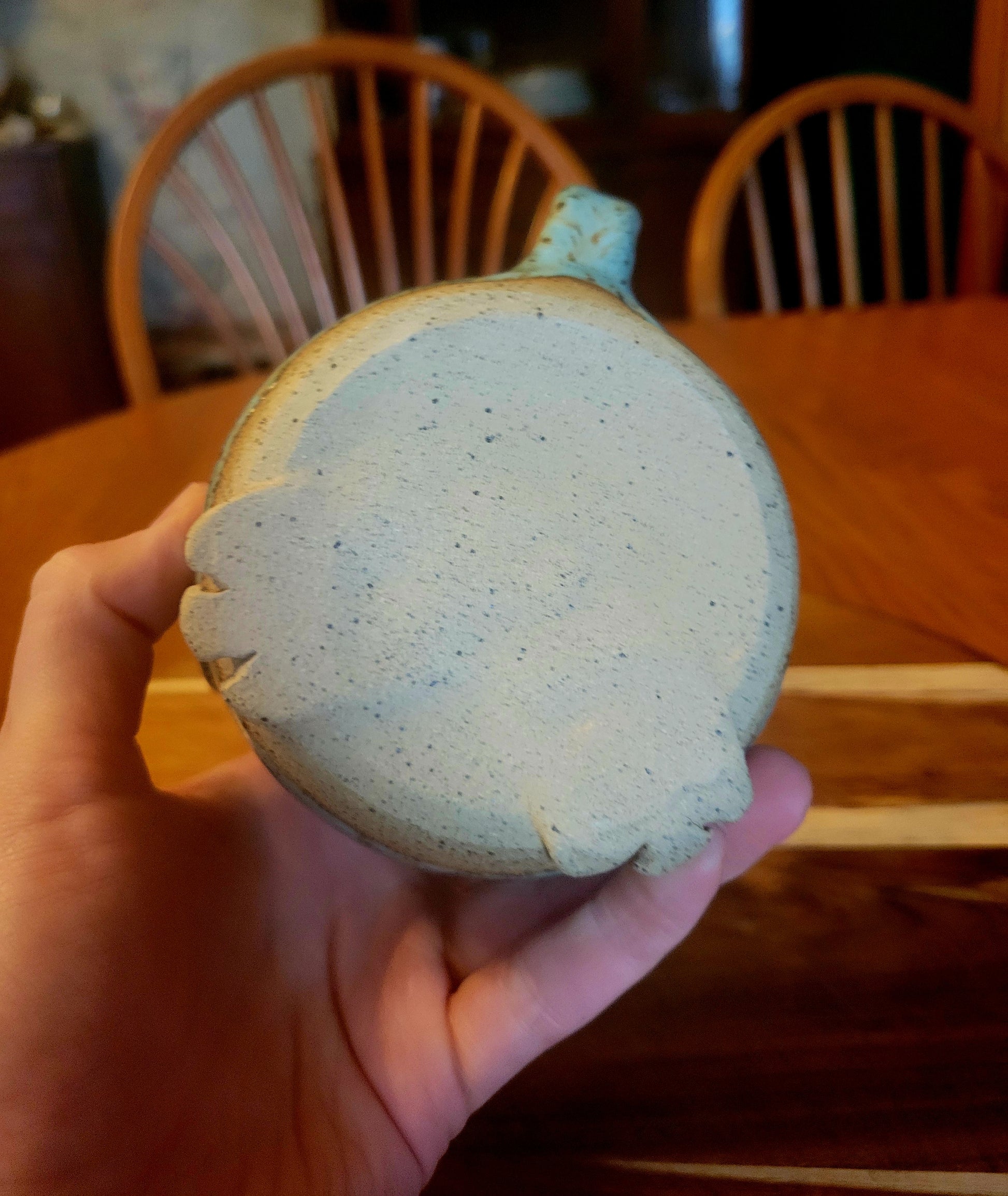 Hand holding a round, speckled ceramic mug with a wooden chair and table in the background.