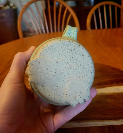 Hand holding a speckled blue and beige mug on a wooden table with chairs in the background.