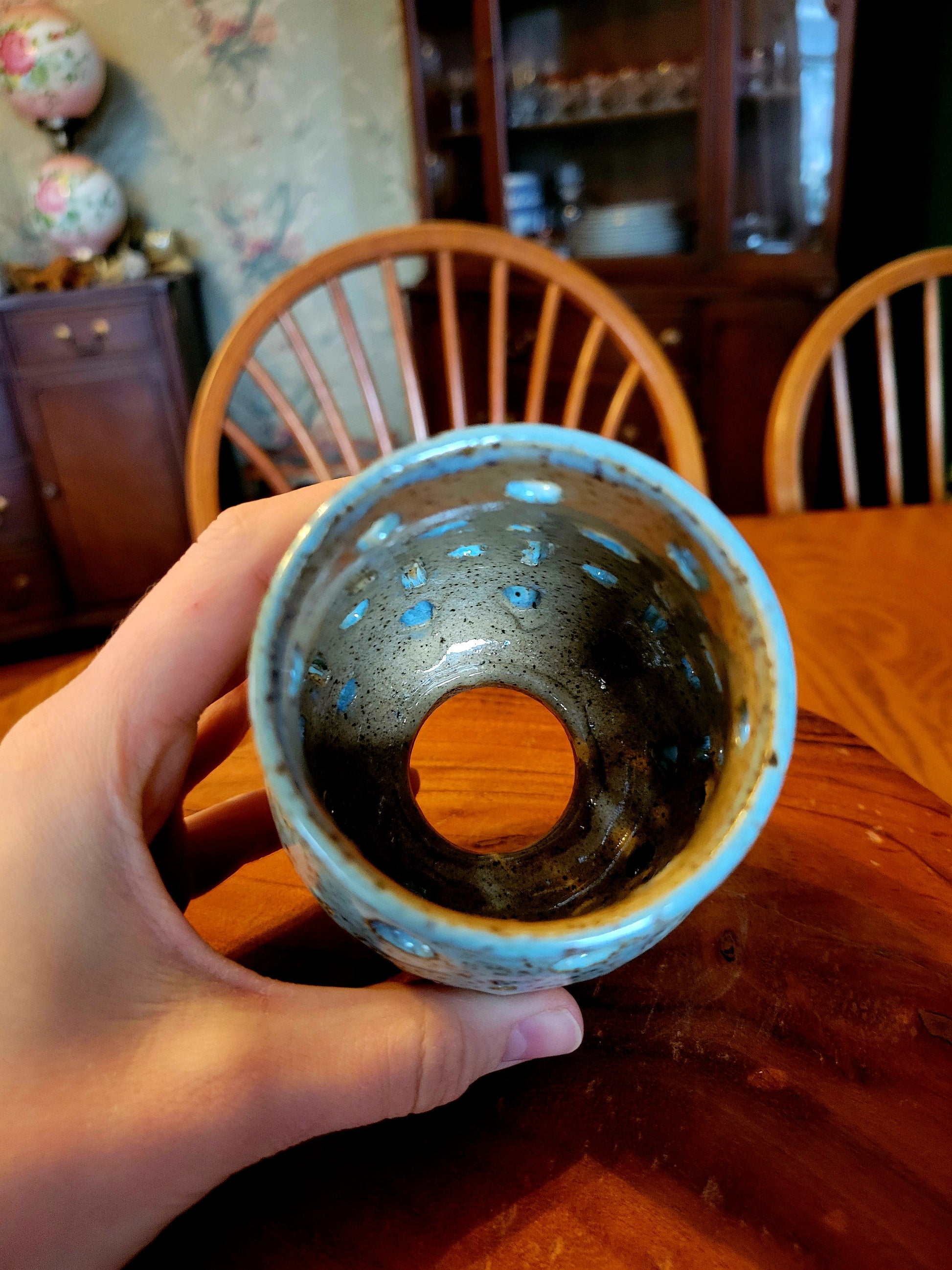 Hand holding a ceramic cup with a textured interior against a wooden chair and table background